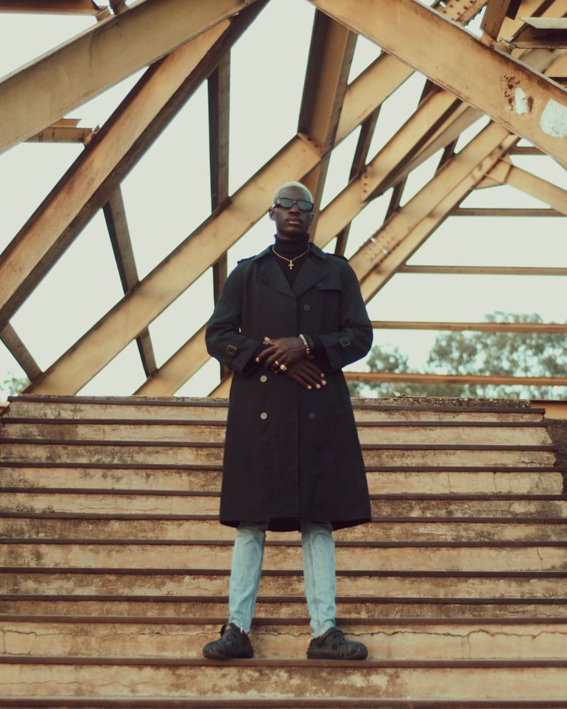 Man in a trench coat posing on a metal staircase under a geometric structure.